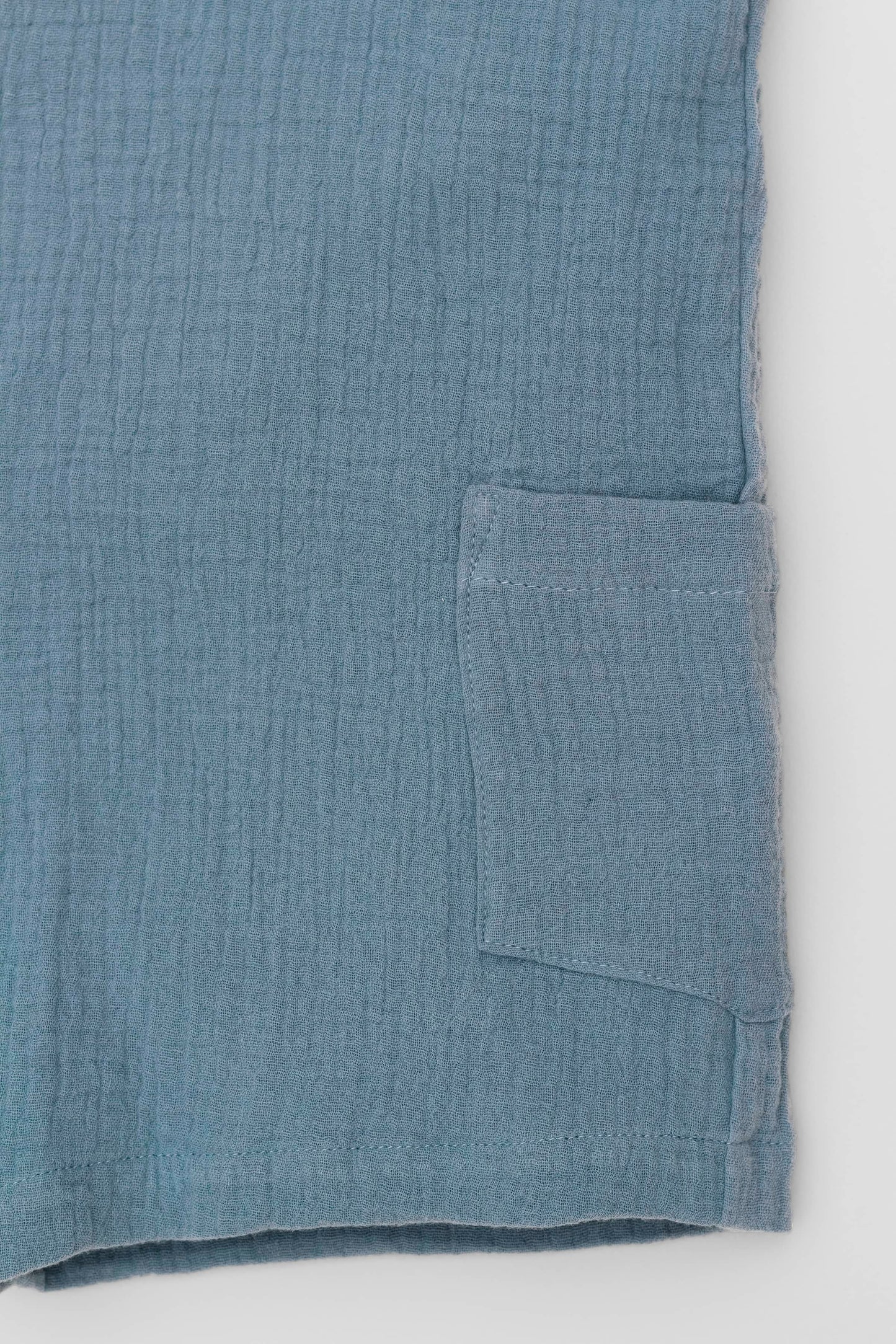 Organic cotton short leg overall in blue muslin. Loose fit and buttoned straps and cargo pockets. Pocket detail